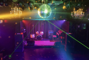 A small stage with a drum set, amplifiers, and a piano is illuminated by colorful lights and a disco ball in a dimly lit venue.